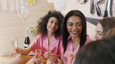 group of multiethnic female friends dressed in pink silk nightdresses talking while toasting with champagne glasses