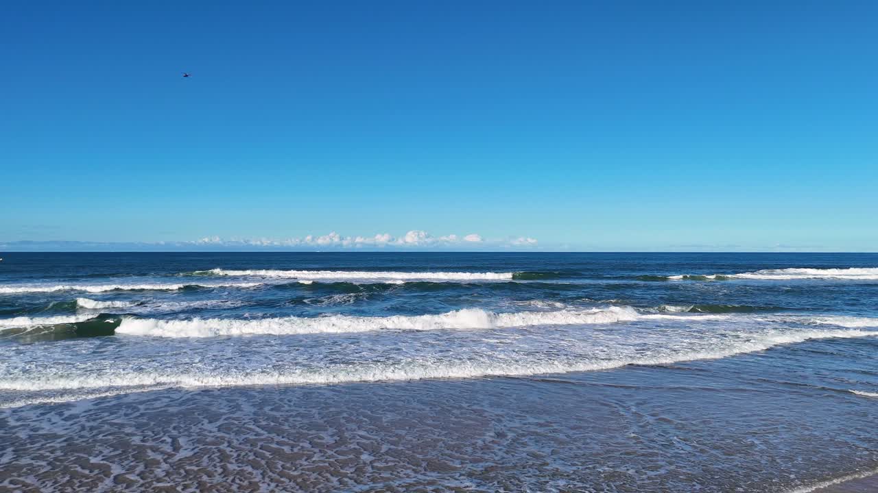 Aerial footage of ocean waves and sandy beach under clear skies at Gold Coast, Australia, showcasing dynamic surf and coastal beauty