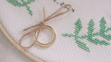 two gold wedding rings tied with twine, placed on a piece of embroidered fabric with green leaf patterns