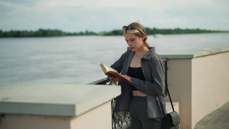 young woman in grey clothing rests her right hand on an iron fence while reading a book, the wind softly blows her hair and clothing as she reads, with trees and riverfront visible in the distance