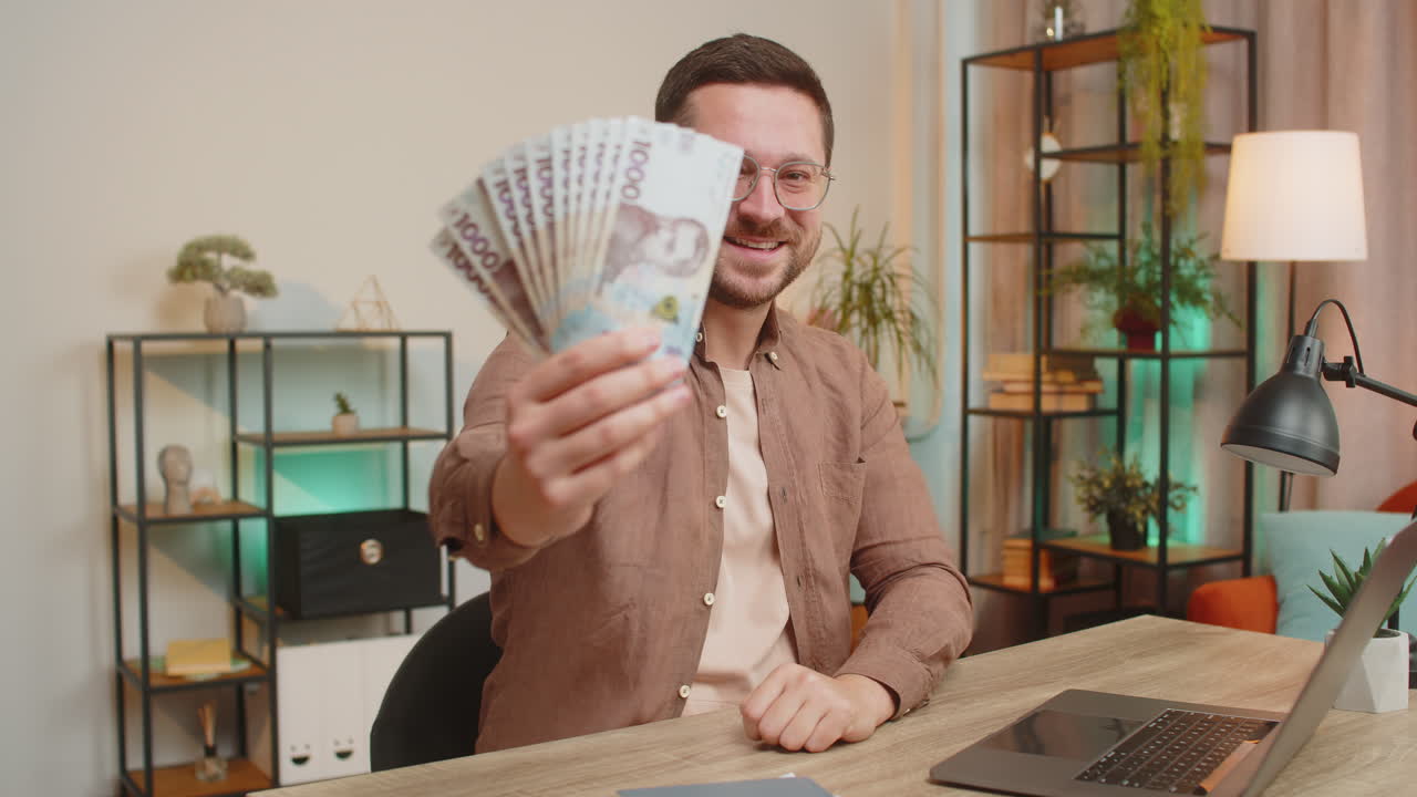 Businessman freelancer holding fan of hryvnias cash showing thumbs up sitting at home office table