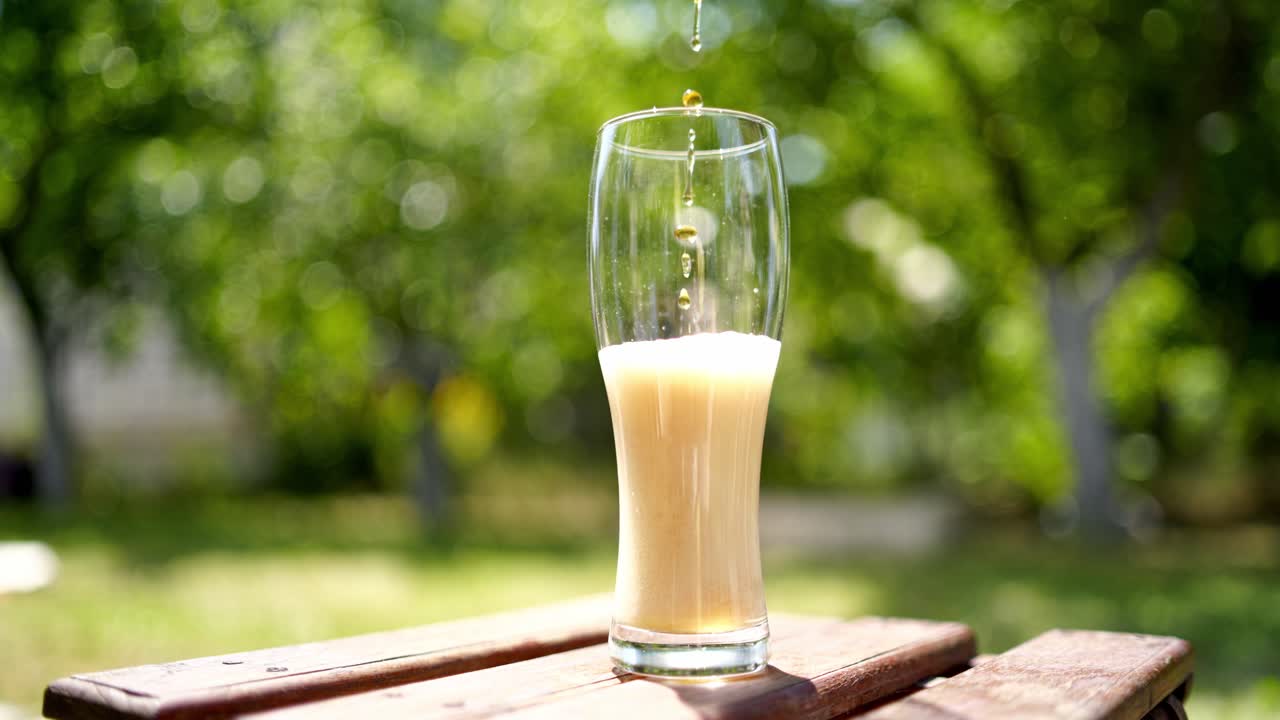 Process of pouring beer to a tall glass on a wooden table. Beer is pouring from the top into the glass with forming wavesfrom the foam. Close-up.