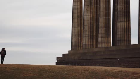 Traveler-walking-next-to-giant-pillars,-Edinburgh,-Scotland,-cloudy,-static-shot