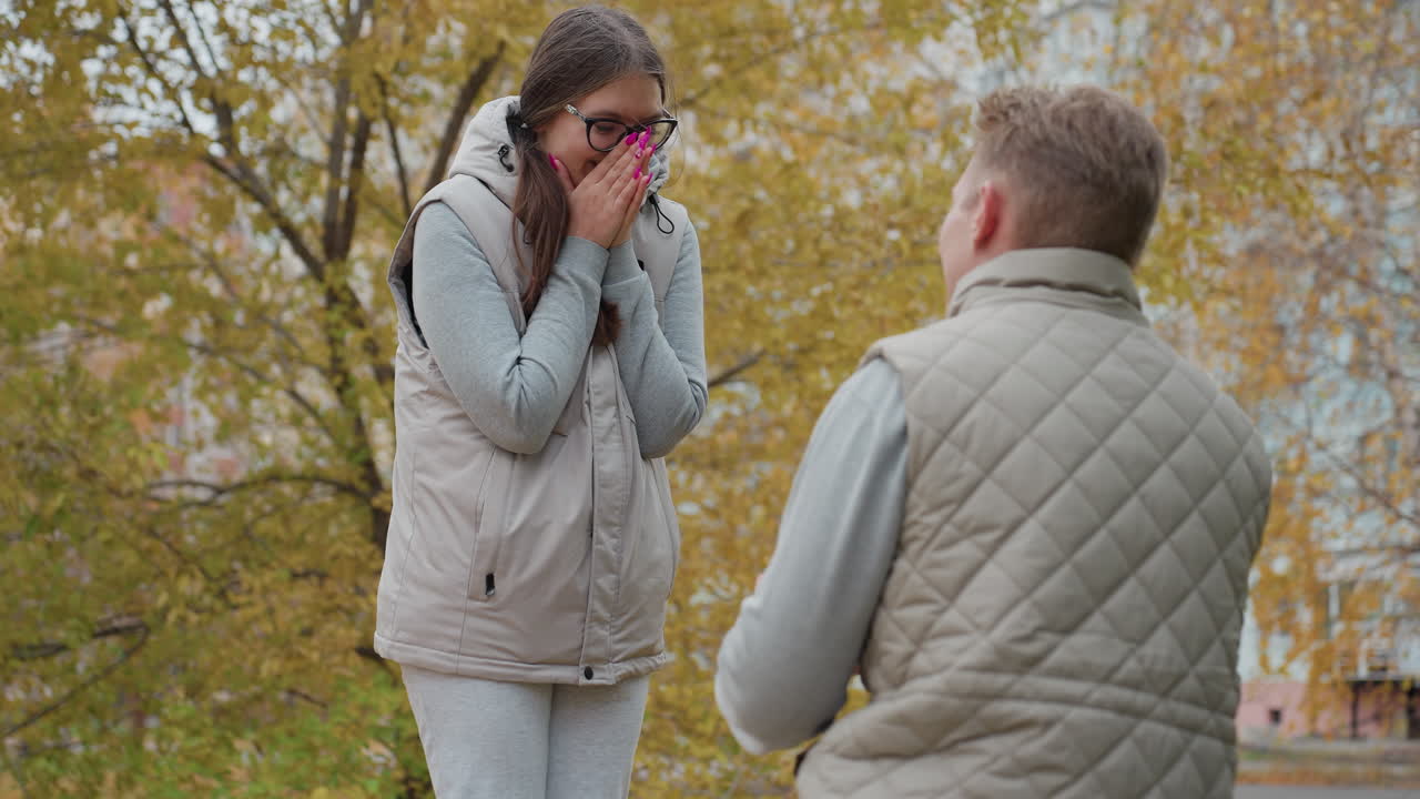 Close up of young man kneeling to propose to girlfriend with painted nails who smiles and nods in agreement while covering her mouth as people walk in background near building