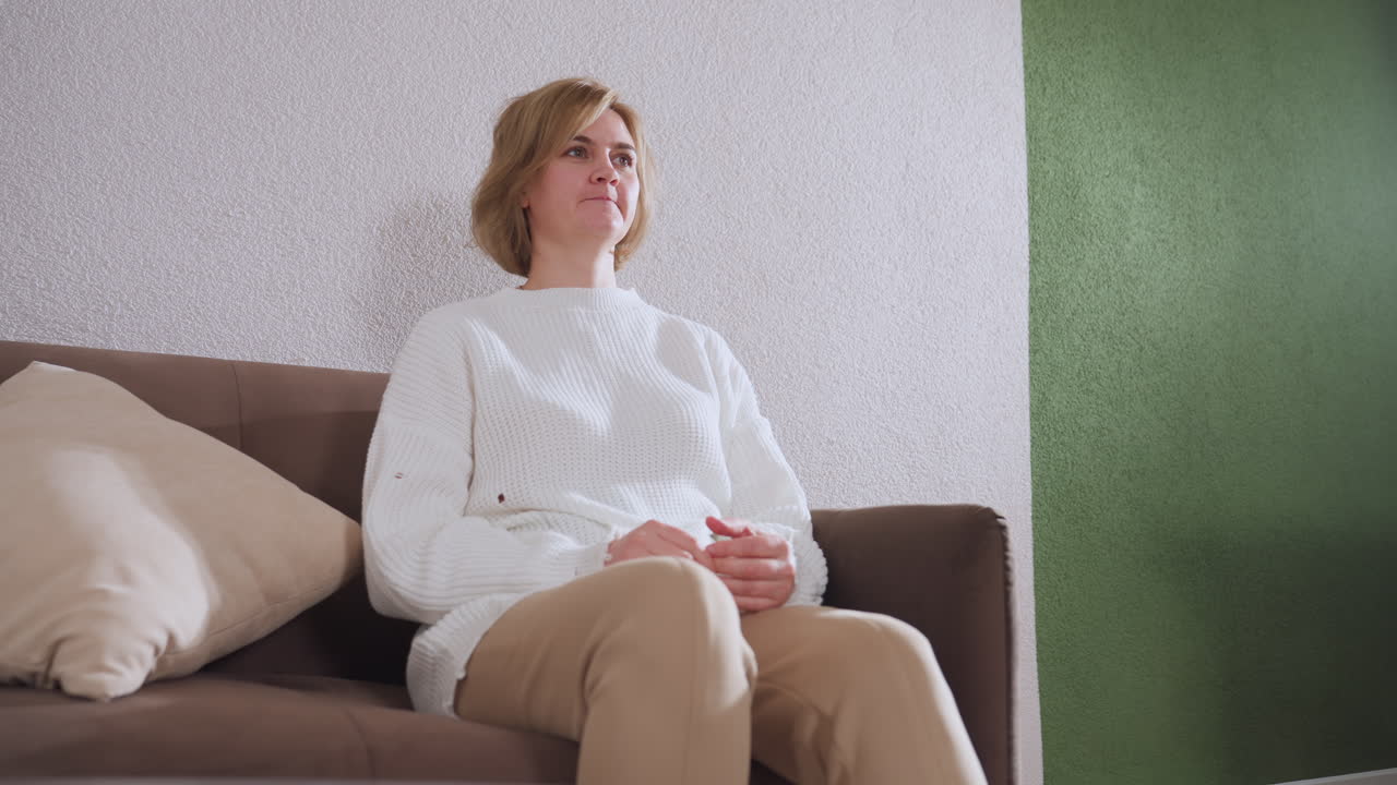 Guest speaking to therapist seated on comfortable couch in cozy therapy room, demonstrating hand gestures while therapist takes notes, cushion behind, pen pot and clock on table, slippers visible