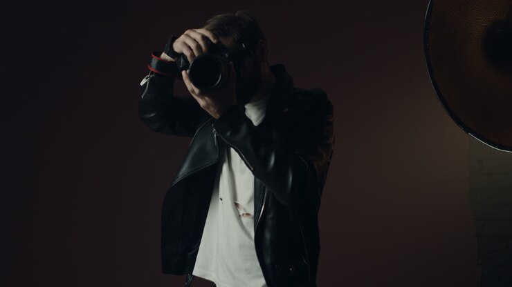 Stylish caucasian man photographer in black lather jacket taking photo with a camera and smiling to the camera in dark studio