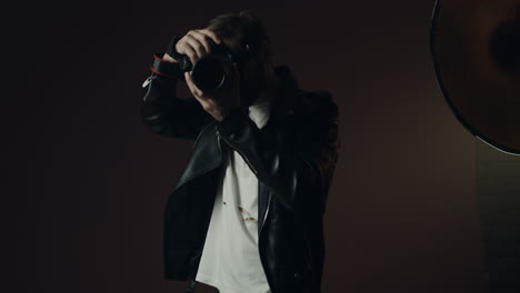 elegante fotógrafo caucásico con chaqueta de espuma negra tomando fotos con una cámara y sonriendo a la cámara en un estudio oscuro
