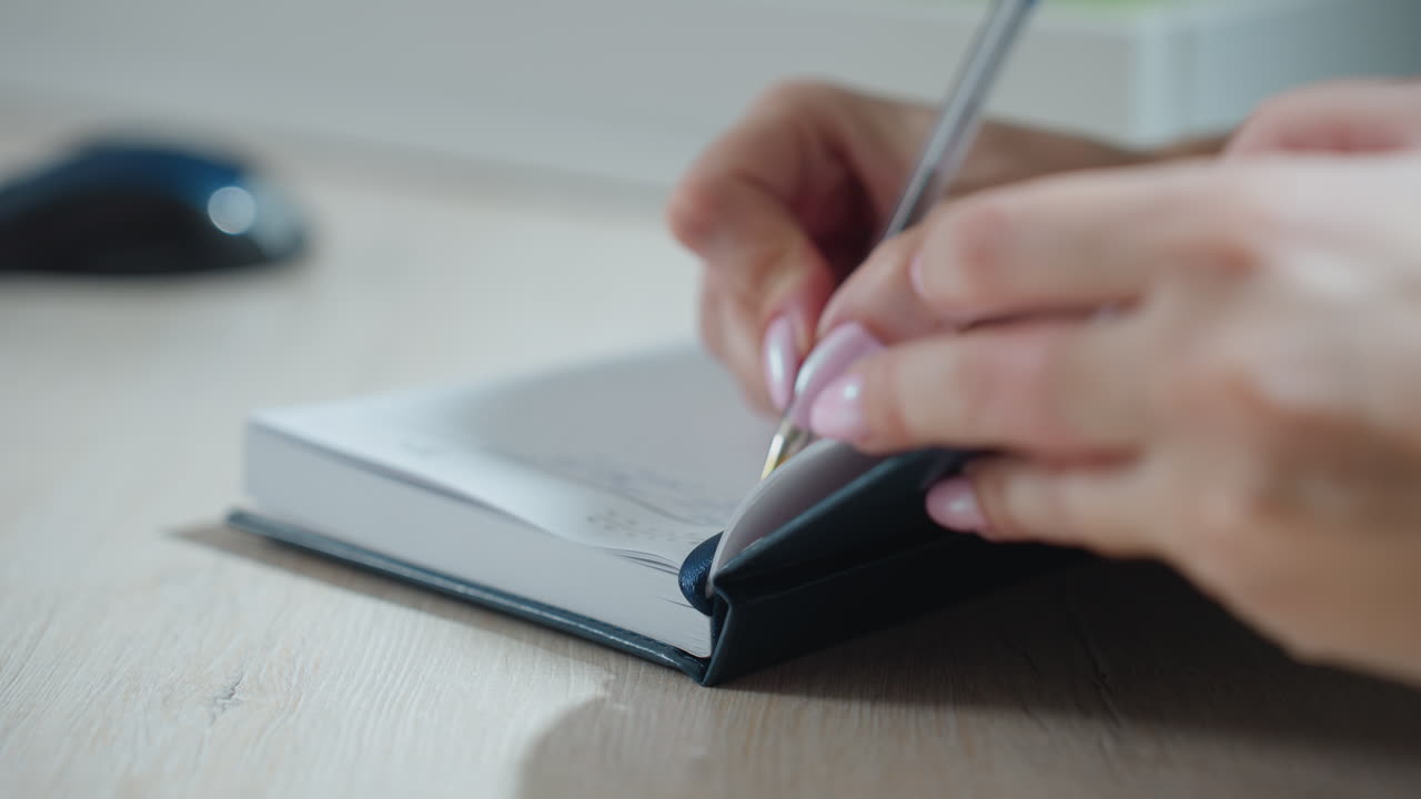 close up of girl writing in notebook with polished pink nails on wooden desk, pen gliding on paper with focused hand movement and soft blur of office environment and objects in background