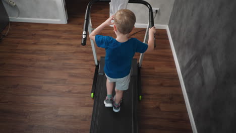 overhead back view of young kid walking on treadmill indoors, rubbing eyes with one hand while holding treadmill handle with other, towel draped on panel, wooden floor and textured wall visible