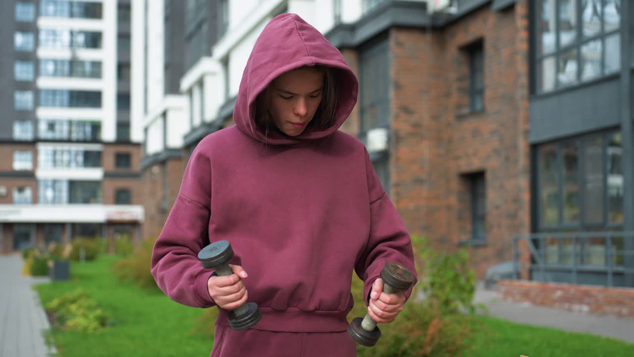Young workout enthusiast in wine hoodie stands outdoors in modern urban courtyard, head bowed in concentration while balancing dumbbells in both hands, embodying strength, and dedication to fitness