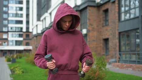 young workout enthusiast in wine hoodie stands outdoors in modern urban courtyard, head bowed in concentration while balancing dumbbells in both hands, embodying strength, and dedication to fitness