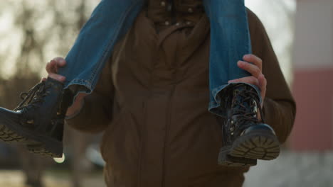 a child sits on a man's neck, showing only their legs. the child is wearing blue jeans and black boots, with the man holding onto their legs