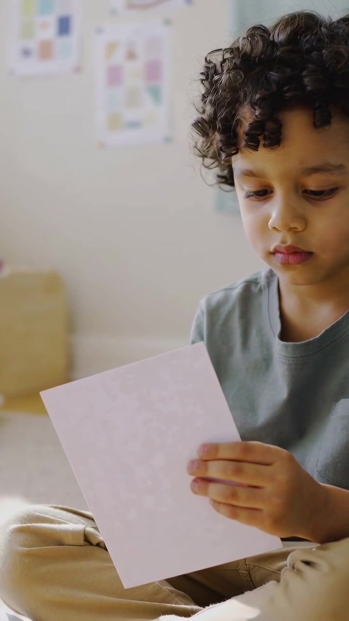 Vertical video: Raising child in green tee trying to read white sheet at home, adjusting grip