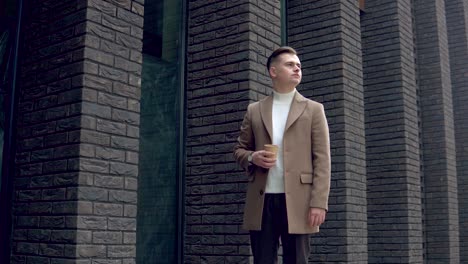 handsome businessman in brown coat with coffee near the office looking at the camera