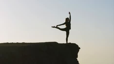woman practicing yoga or dance pose on cliff edge at sunrise or sunset