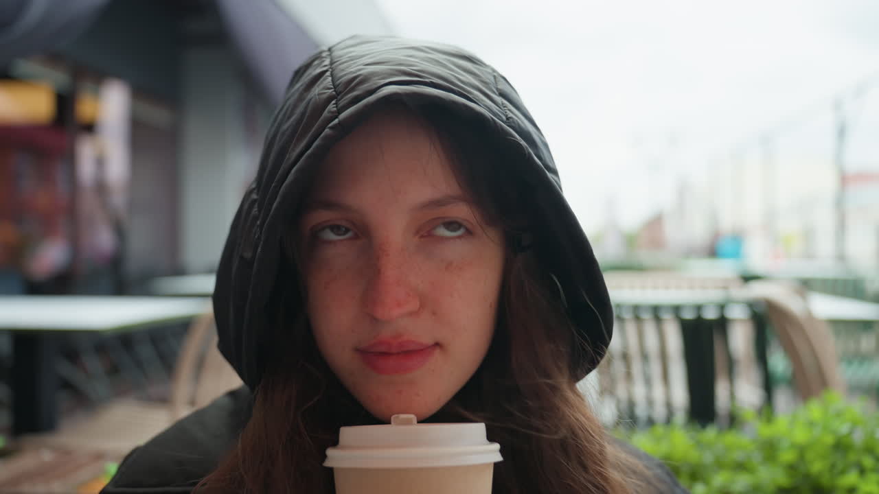 フード付きジャケットを着た女性が、テイクアウトのコーヒーカップを持っているクローズアップ写真。雨の日の都市の市場のテラスでのクローズアップポートレート、ソフトなボケ味の背景、そばかすと繊細な表情、内省的な視線、映画のような雰囲気