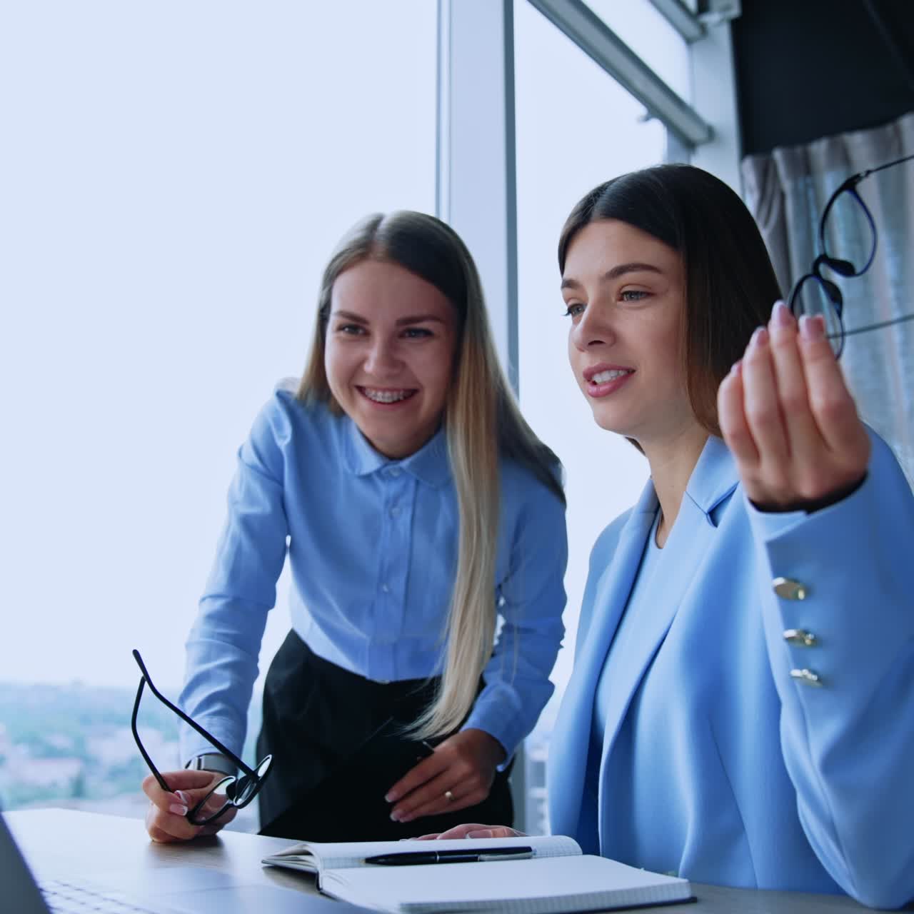 Happy resilient female employees in modern office. Ladies look at laptop on the desk, discussing what they see an laughing cheerfully. Low angle view