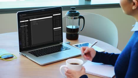 Caucasian-woman-sitting-at-table-watching-coding-data-processing-on-laptop-screen-and-making-notes