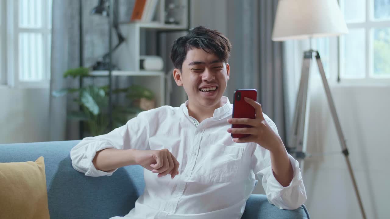 Smiling Asian Man Having Video Call On Smartphone While Lying On Sofa In The Living Room