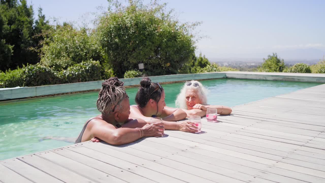 Drei Freundinnen in Badekleidung lehnen am Pool, die Frau in der Mitte gestikuliert einer Freundin zu und hebt ihre rosa Sonnenbrille.