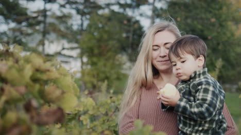 a young mother holds a baby in her arms, he eats a large apple. in the foreground is a vine, in the back is a forest