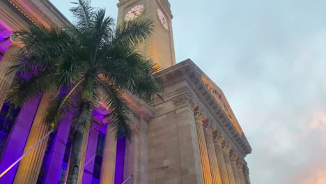 Vista-Panorámica-De-La-Puesta-De-Sol-Naranja-De-La-Torre-Del-Reloj-Del-Ayuntamiento-De-La-Ciudad-De-Brisbane-Con-Luces-Violetas-Que-Iluminan-Las-Palmeras