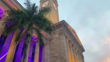 vista panorámica de la puesta de sol naranja de la torre del reloj del ayuntamiento de la ciudad de brisbane con luces violetas que iluminan las palmeras