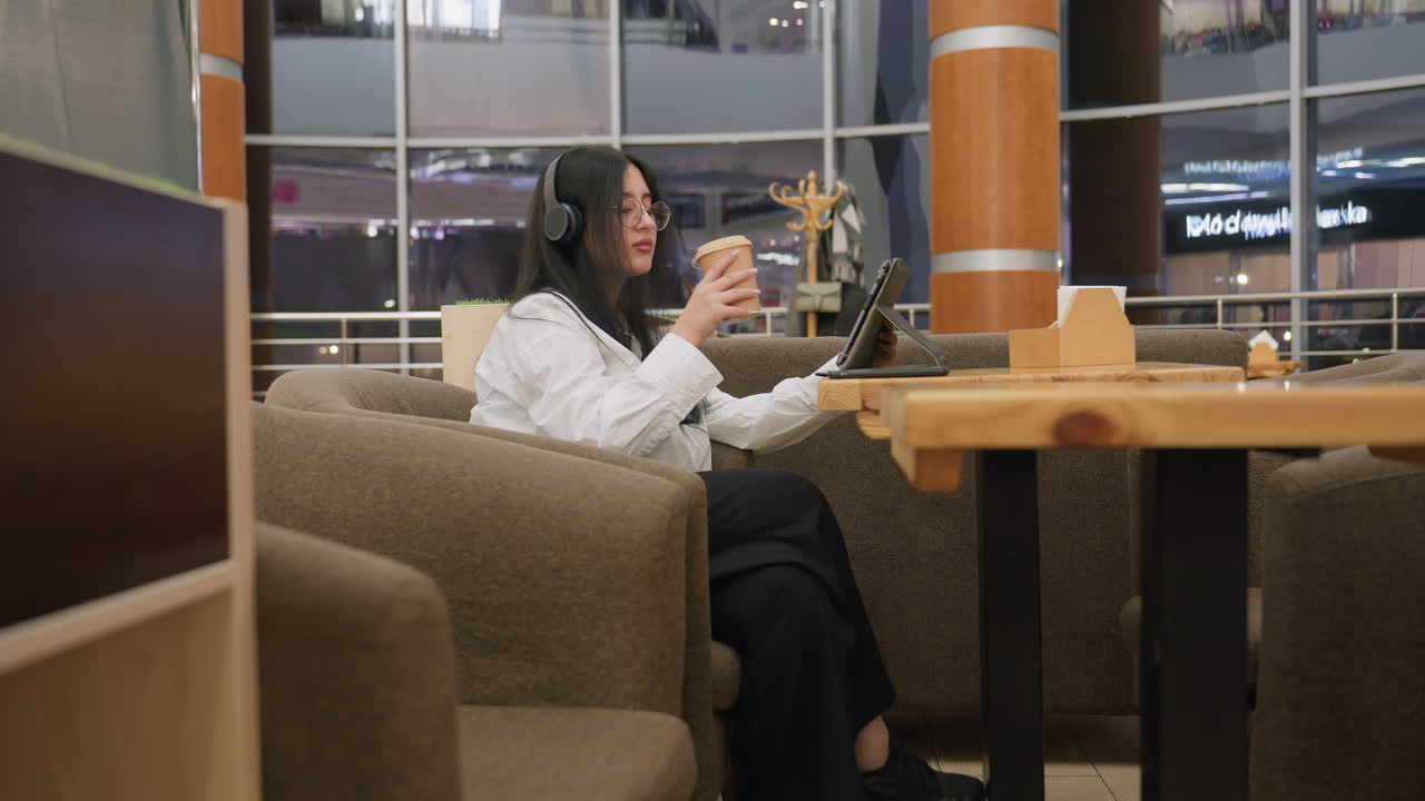 focused woman wearing headphones and glasses places coffee cup on wooden table while working on tablet in cozy cafe with city view background and modern interior during evening hours