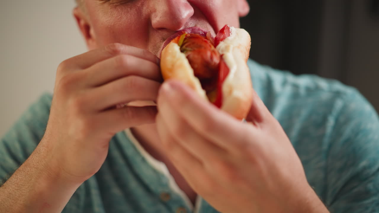 Close up of hungry man eating large sandwich filled with sausage, vegetables, and sauce during lunch, biting with appetite and holding sandwich with both hands, wearing casual blue shirt