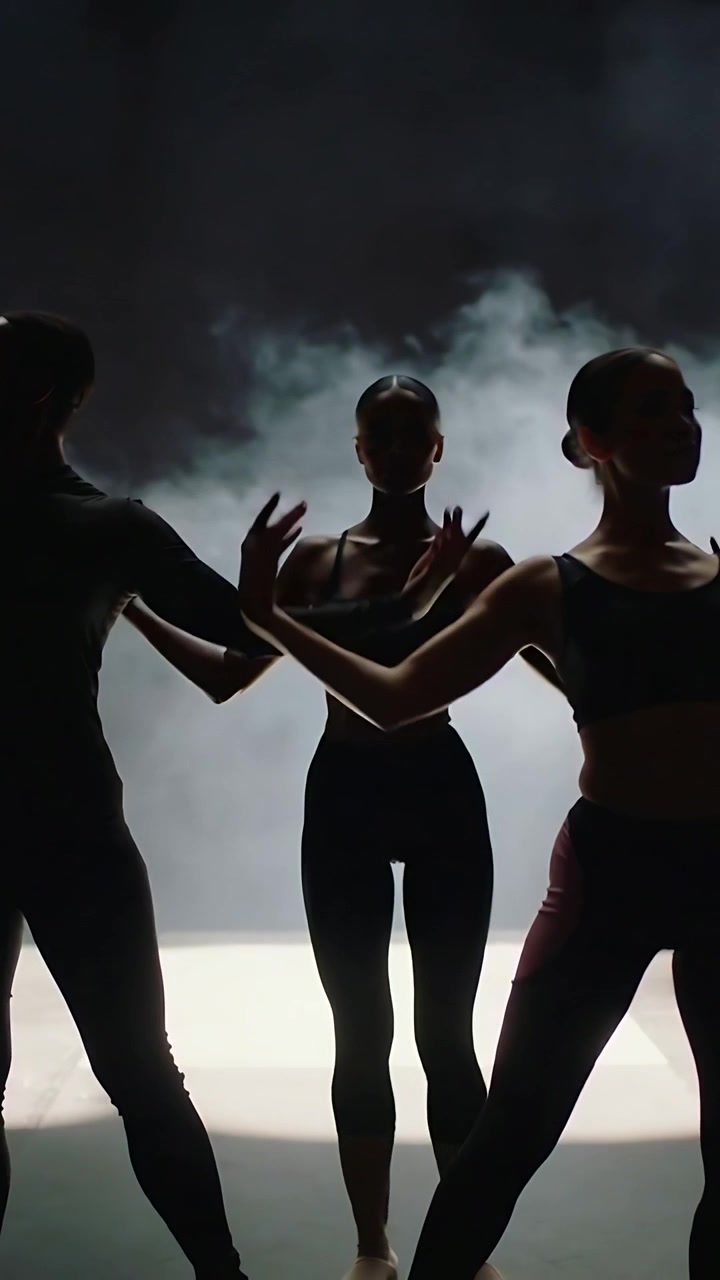 Video verticale: Trio illuminato da controluce, quello centrale in body, che forma figure a strati su un palco nella nebbia