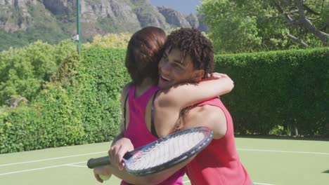 Happy-biracial-couple-with-tennis-rackets-embracing-in-garden-on-sunny-day