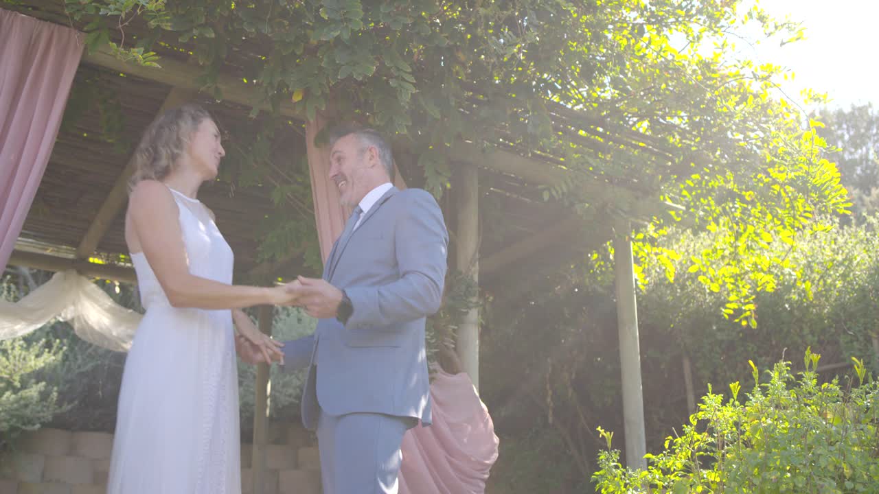 Celebrating couple dancing under pergola, man in suit lifting white-dress woman's hand, embracing
