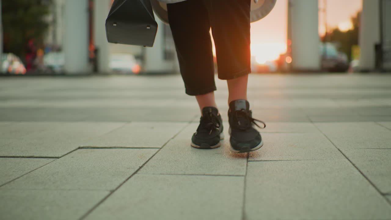 leg view of woman walking confidently on city pavement wearing black canvas sneakers and carrying black handbag with soft sunset glow in background