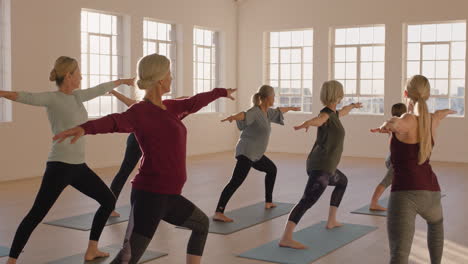 yoga class of healthy mature women practicing warrior pose enjoying morning physical fitness exercise workout in studio at sunrise