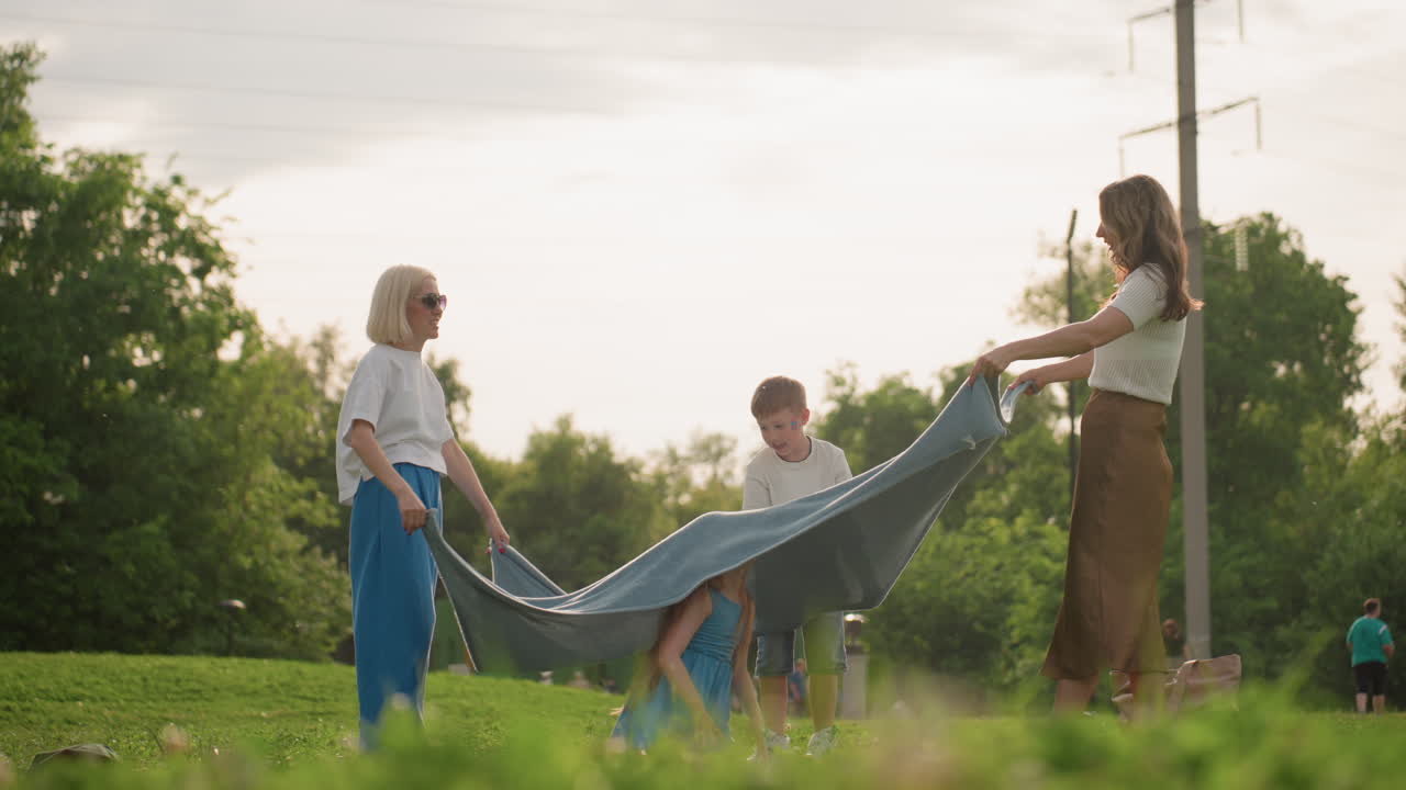 Nurturing women and girl in middle playfully swaying cloth over head in sunny park, green grass background, laughter and movement, casual summer clothes, joyful family connection and outdoor bonding