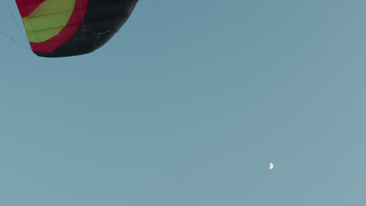 Colorful paraglider canopy drifting against vast twilight sky with half moon visible above serene landscape, capturing gentle motion and contrast of glider silhouette against soft dusk light