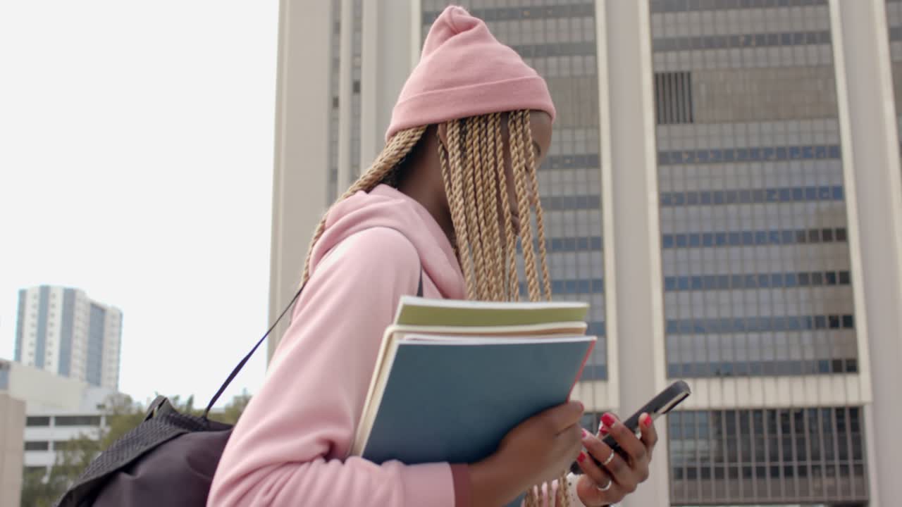 Notification pinging African American woman scanning phone while walking plaza carrying notebooks