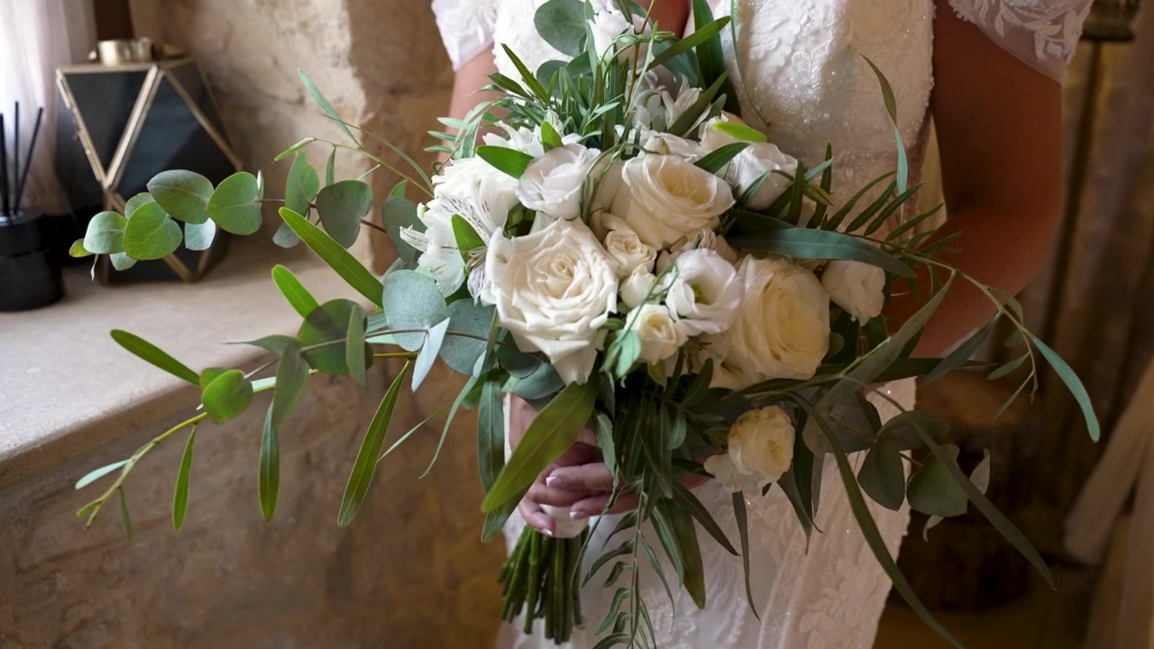 Una sposa regge un bellissimo bouquet di rose bianche e verde in una stanza incantevole e dalla luce soffusa. La composizione aggiunge un tocco di eleganza all'atmosfera nuziale.