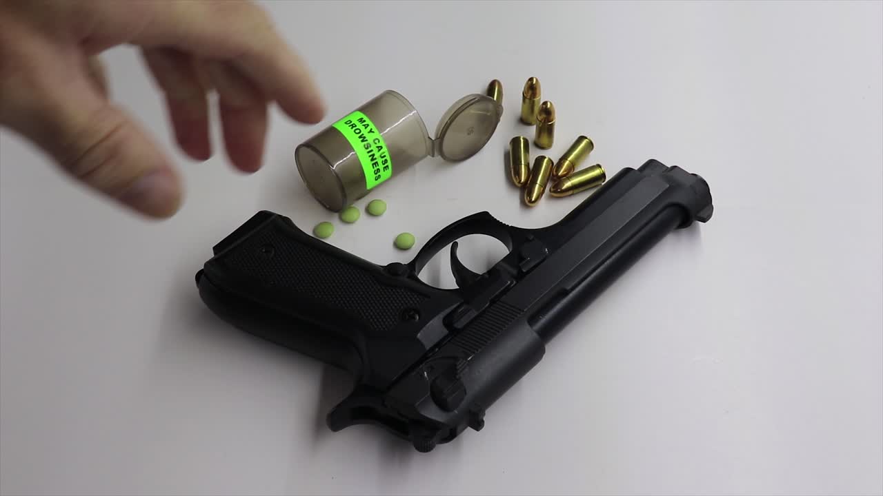 persona recogiendo un arma de fuego negra con una botella de medicamentos de pastillas vacías con un signo de somnolencia de precaución y pastillas al lado del arma de fuego rodeada de algunas balas de 9 mm