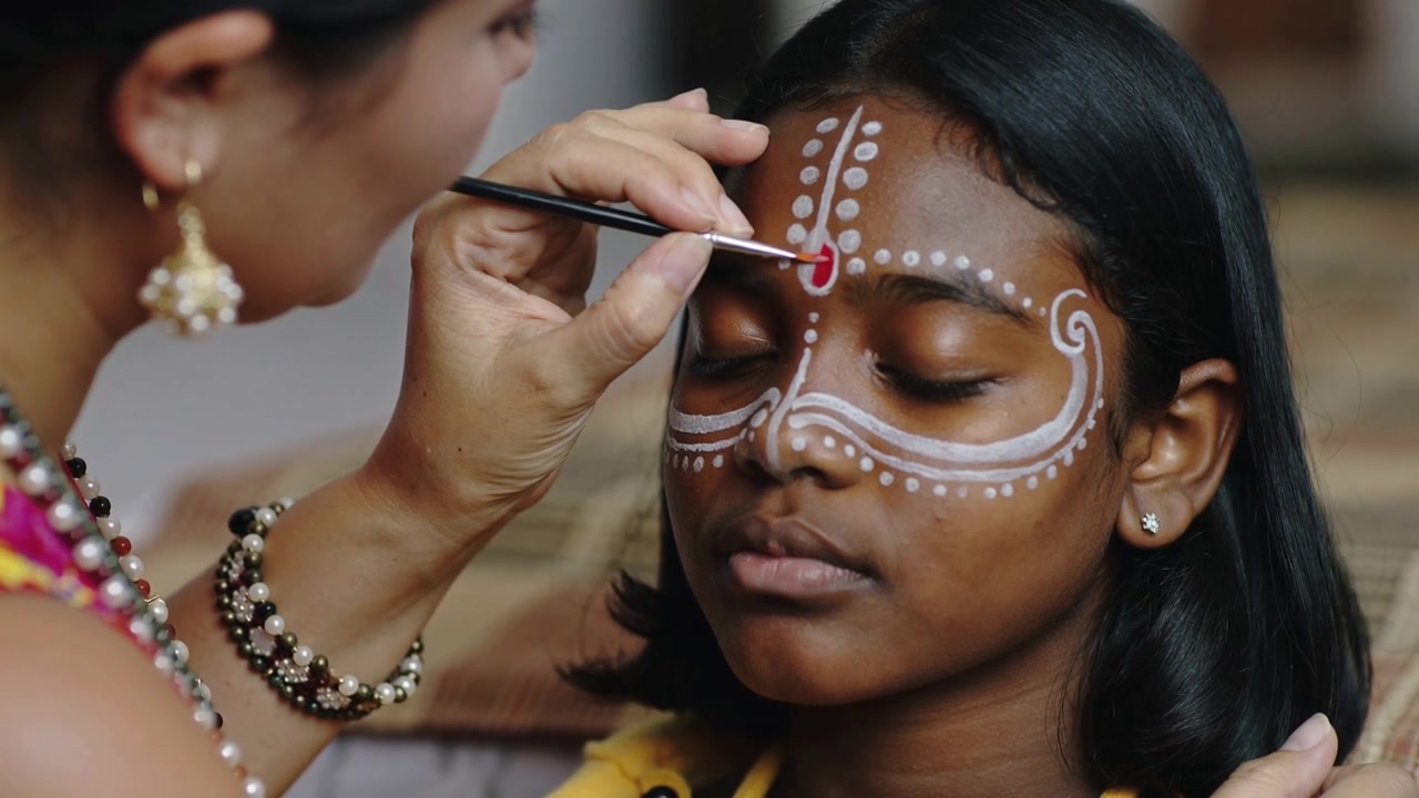 Bringing brush painter steadying girl, painting red teardrop and white-dot mask on forehead at home