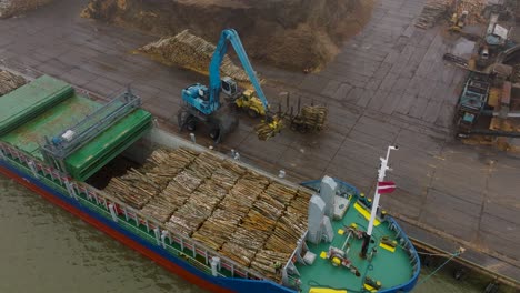 aerial establishing view of wood terminal crane loading timber into the cargo ship, port of liepaja , lumber log export, overcast day with fog and mist, wide birdseye drone shot