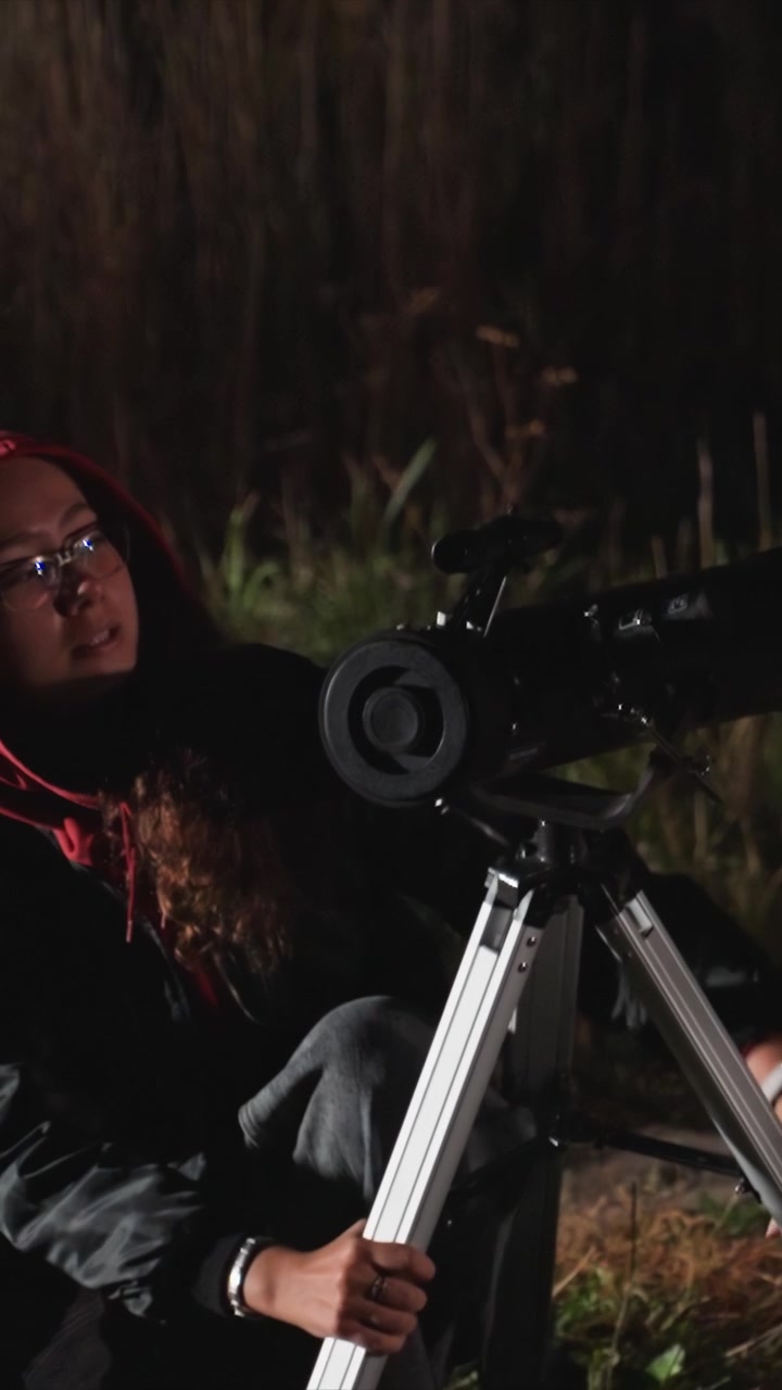 Woman wearing glasses adjusts tripod for large optical. Woman wears multiple rings on left hand as she works. Setting includes multi story building with illuminated panes