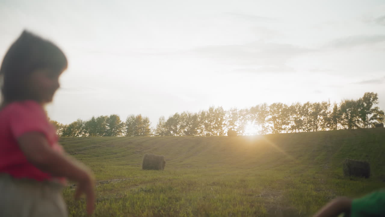 분홍색 옷을 입은 어린 소녀는 녹색 셔츠를 입은 동생에게 놀이롭게 풀을 던지고, 동생은 웃으며, 황금빛 해가 지는 동안 시골 에서 야외에서 재미있는 순간을 즐니다.
