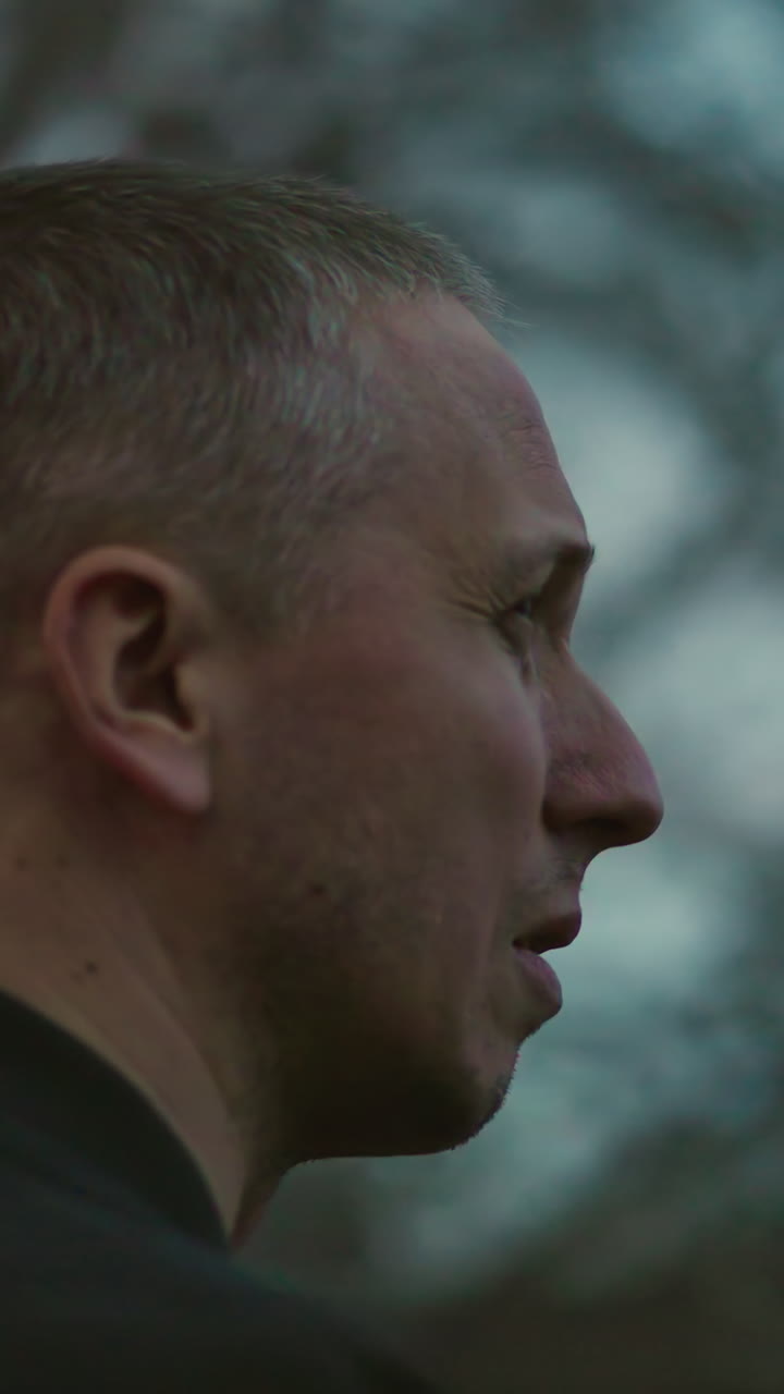 A man in a green jacket smokes a cigarette outdoors at dusk, with smoke visibly escaping his nose and mouth