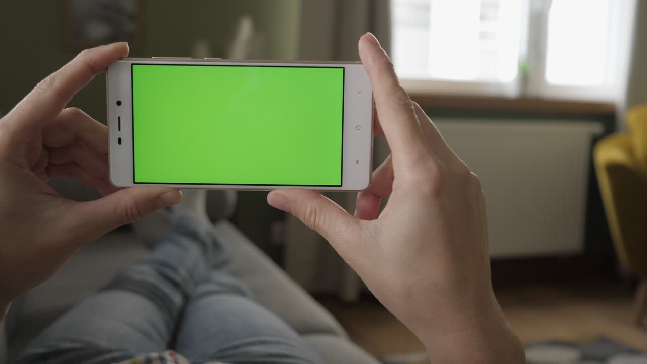 젊은 여성이 수평 모드에서 녹색 화면 스마트폰과 함께 소파에 누워있는 집. 터치 스크린 휴대전화를 사용하는 소녀. 스마트폰을 사용하는 소녀, 인터넷 브라우징, 비디오 콘텐츠, 블로그, pov.