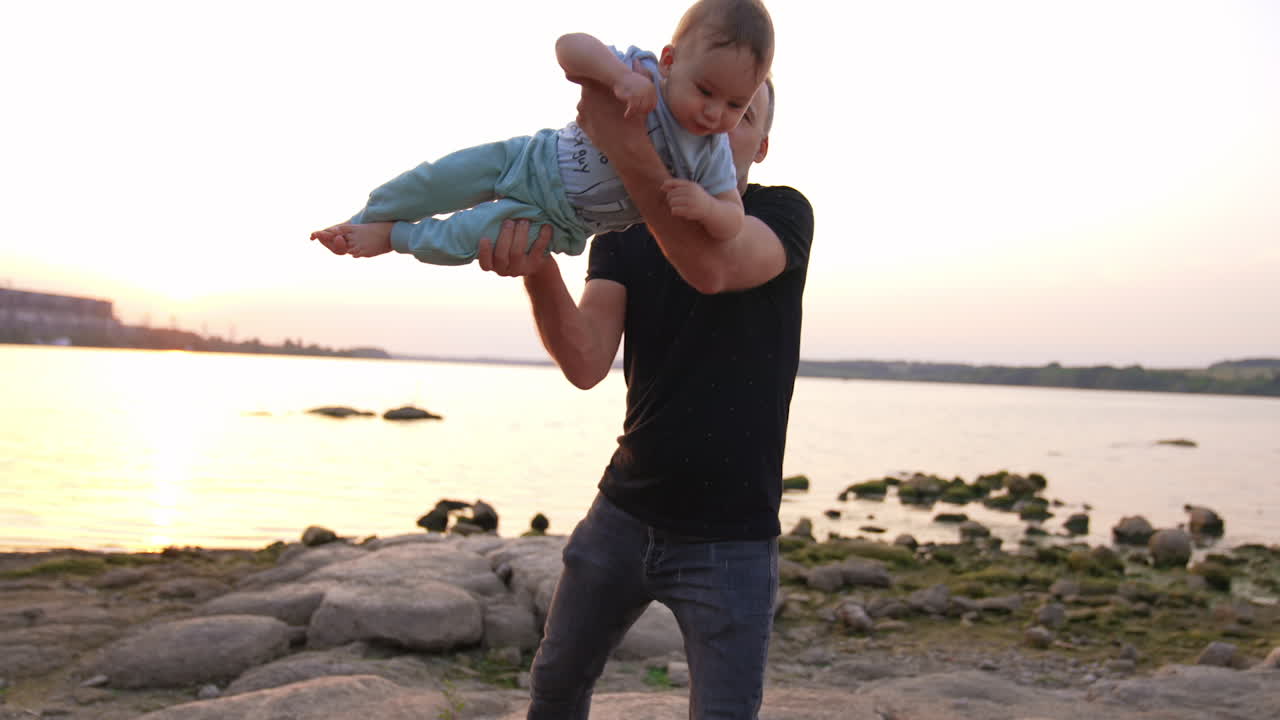 Beautiful little baby flying in the hands of his careful dad. Family spending time at the river bank in the end of the day.