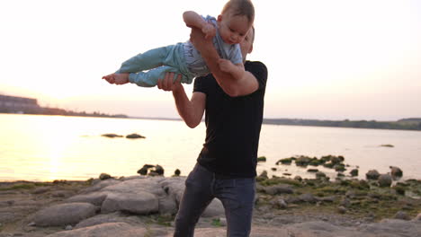 beautiful little baby flying in the hands of his careful dad. family spending time at the river bank in the end of the day.