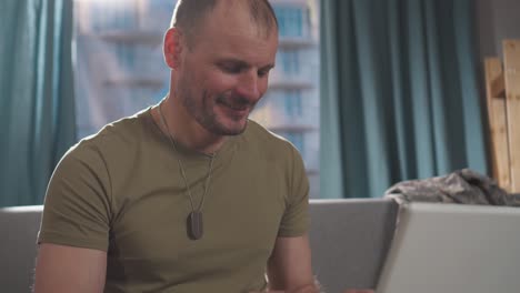 portrait of a positive man a professional military man in uniform ,sitting on a sofa with a living room at home and using a notebook close up