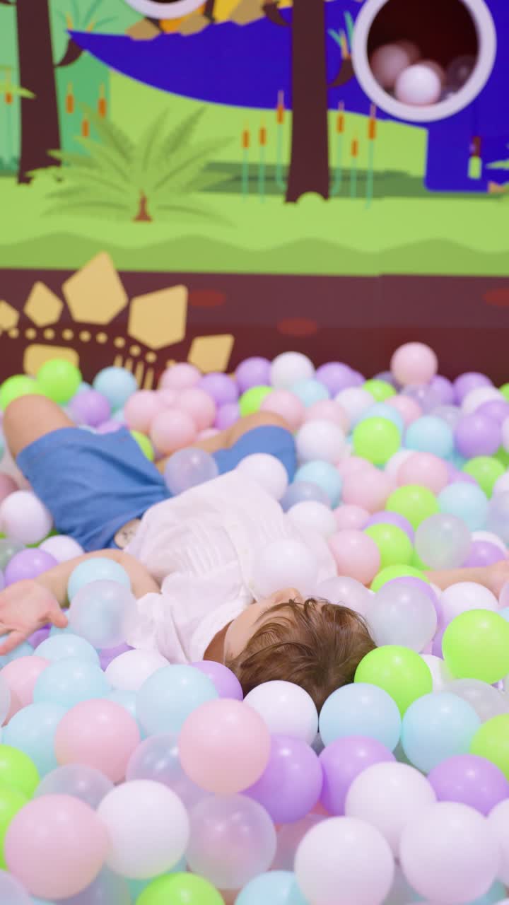 A cheerful 5 year old girl lies on her back, playing and throwing colorful plastic balls in a play pit with dinosaur themed walls