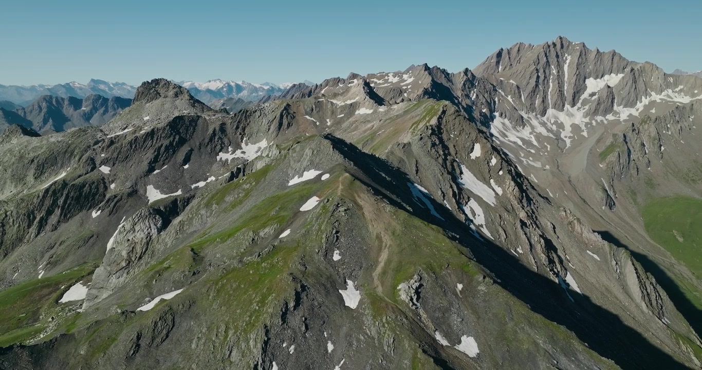 Drone che orbita attorno a una spettacolare vetta alpina svizzera, rivelando catene montuose a strati, creste di granito frastagliate, formazioni geologiche e cime innevate sotto un cielo estivo limpido e azzurro.